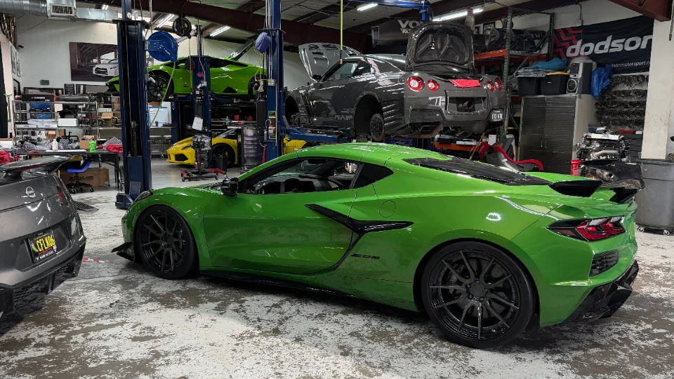 A green Corvette C8 Z06 parked in a performance tuning shop with a Nissan GT-R and Lamborghini Huracan on lifts.