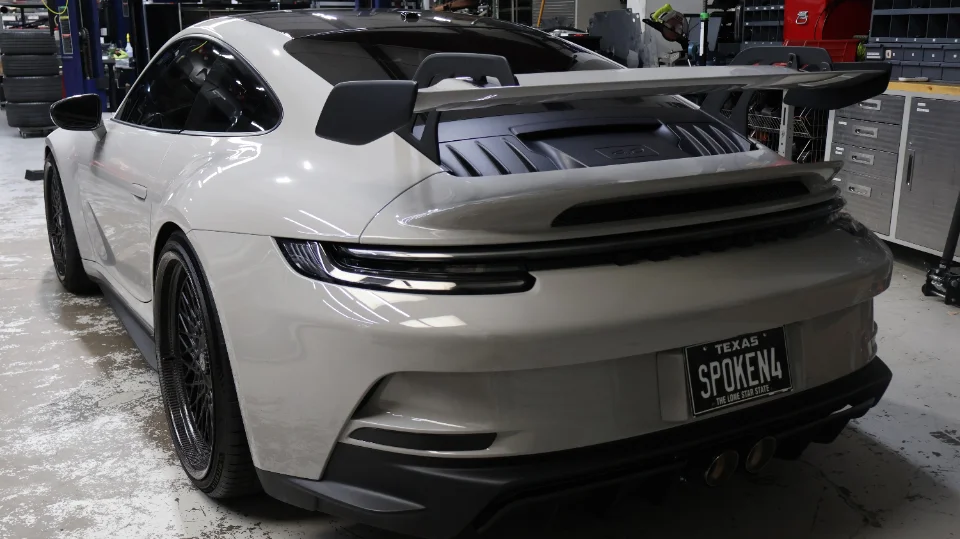 A white Porsche 911 GT3 with a large rear wing and custom Texas license plate parked inside a performance shop.
