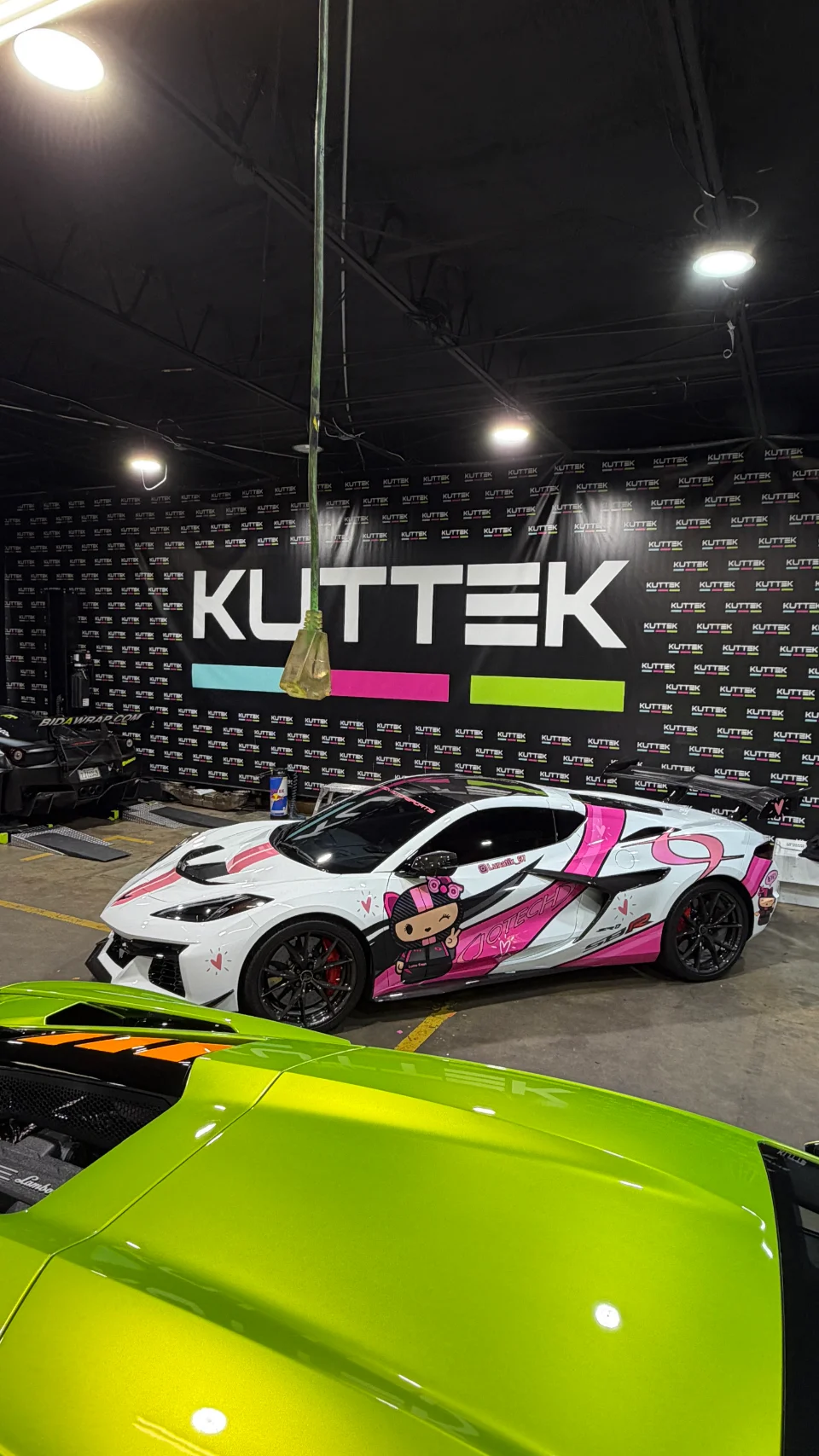 A white Corvette C8 with a custom pink anime wrap and a green Lamborghini parked inside a KUTTEK performance shop.