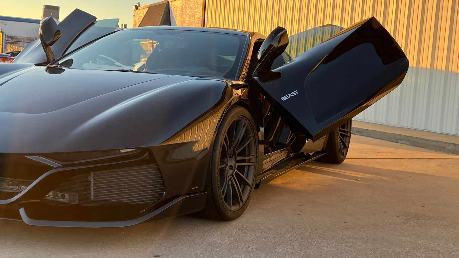 A sleek black sports car with unique upward-opening doors, parked outdoors in golden hour light, featuring 'BEAST' branding.