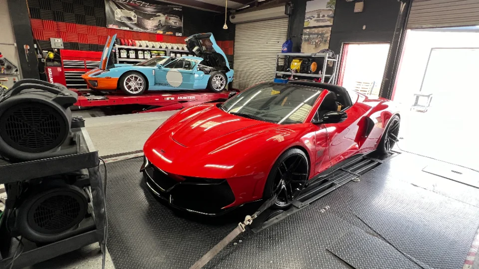 A red supercar strapped to a dynamometer in a performance tuning shop, with a blue Ford GT on a lift in the background.