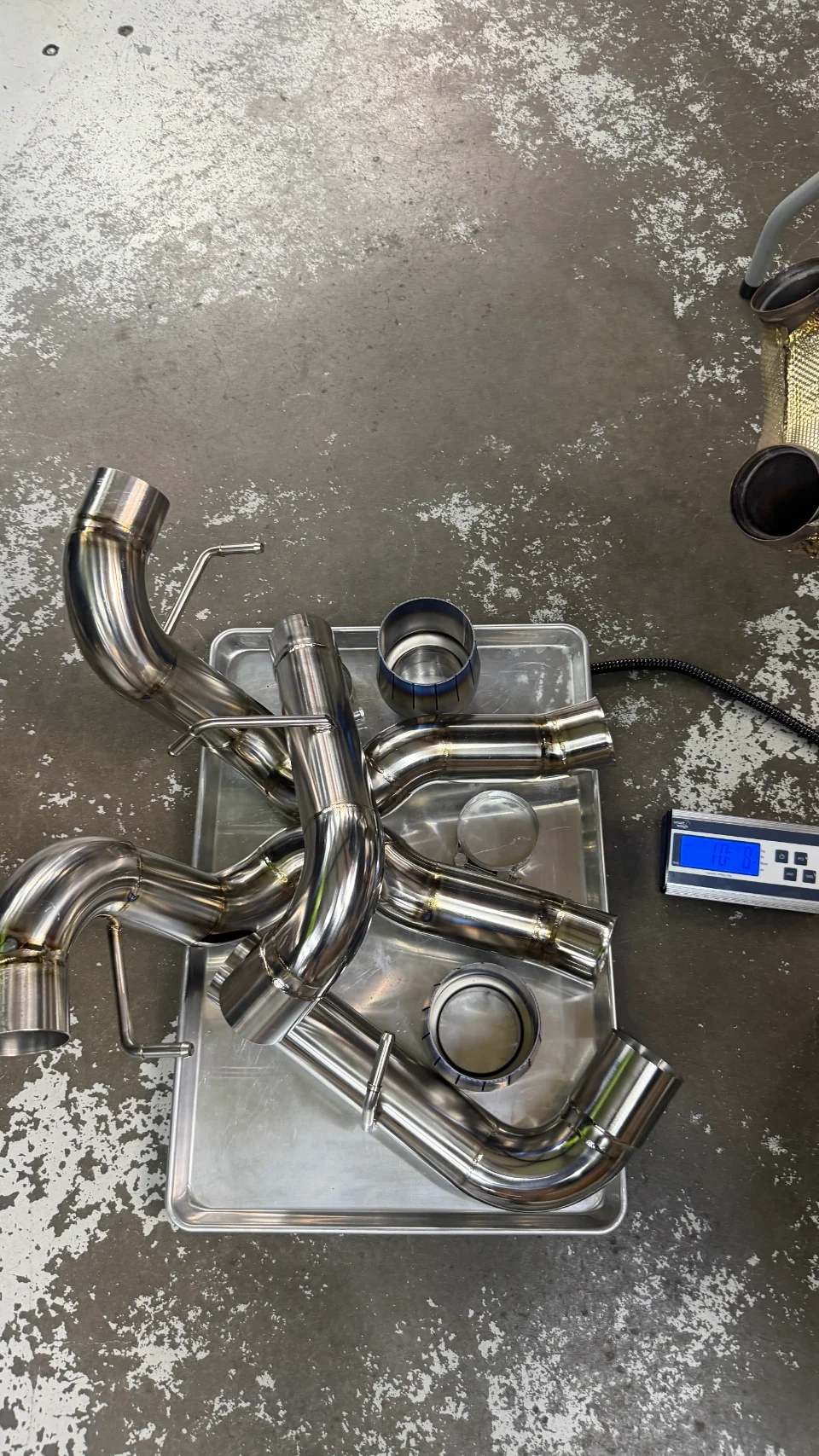 Stainless steel exhaust components laid out on a metal tray next to a digital scale in a performance shop.