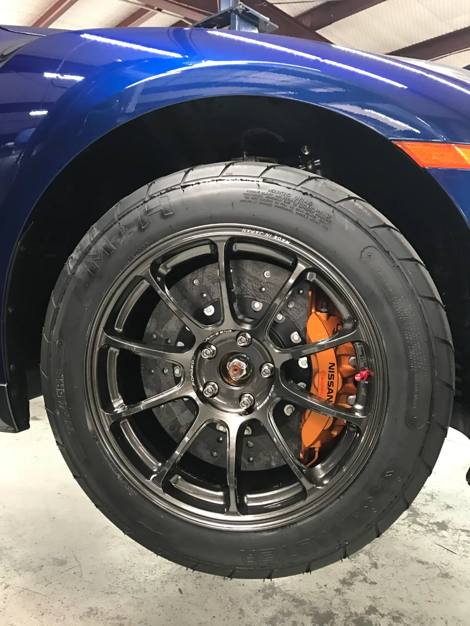 A blue Nissan GT-R's front wheel with dark grey Rays Volk Racing wheels, orange Brembo brake calipers, and a drag radial tire.