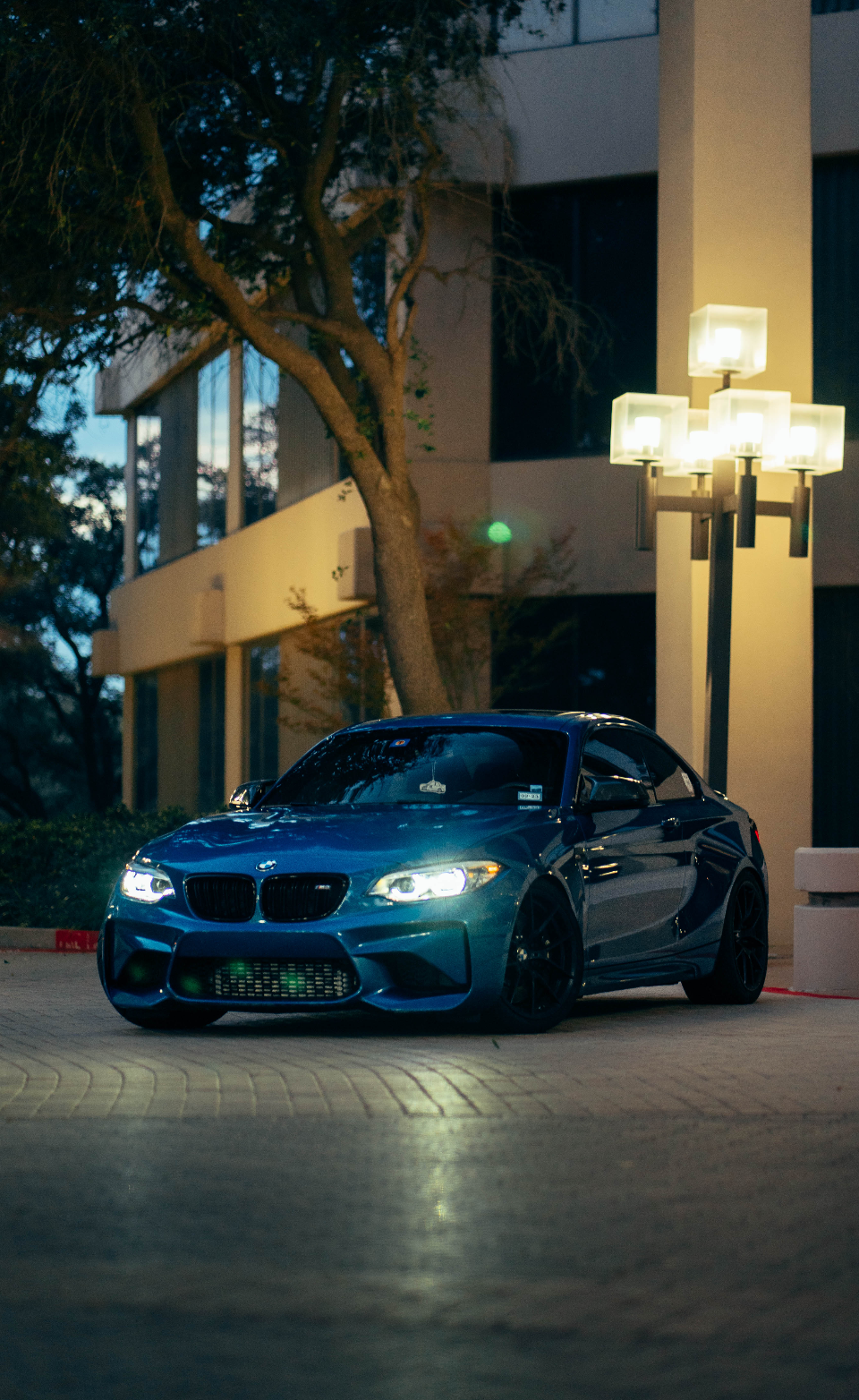 A striking blue BMW M2 coupe parked at night in an urban setting, illuminated by building lights and a street lamp.