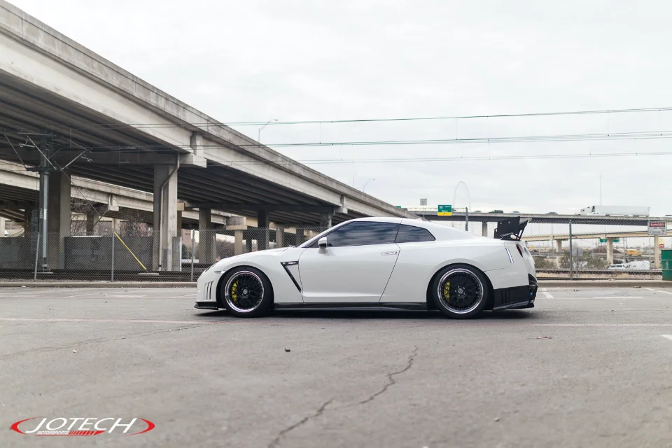 White Nissan GT-R with aftermarket wheels and a large rear wing parked under an urban overpass.