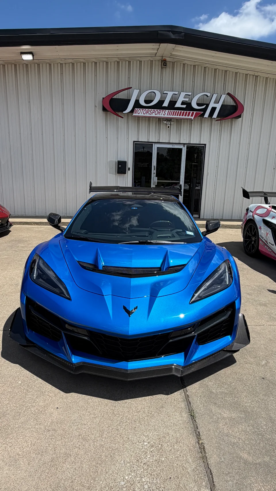 A vibrant blue Chevrolet Corvette Z06 with aggressive carbon fiber aero parked prominently in front of the Jotech Motorsports shop.