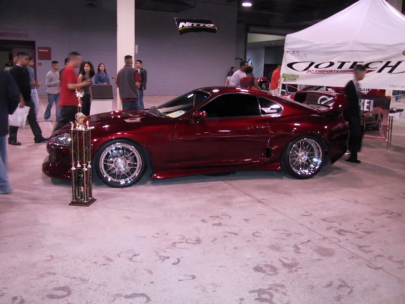 A custom candy apple red Toyota Supra on display at a car show, with a trophy next to its front wheel and people in the background.