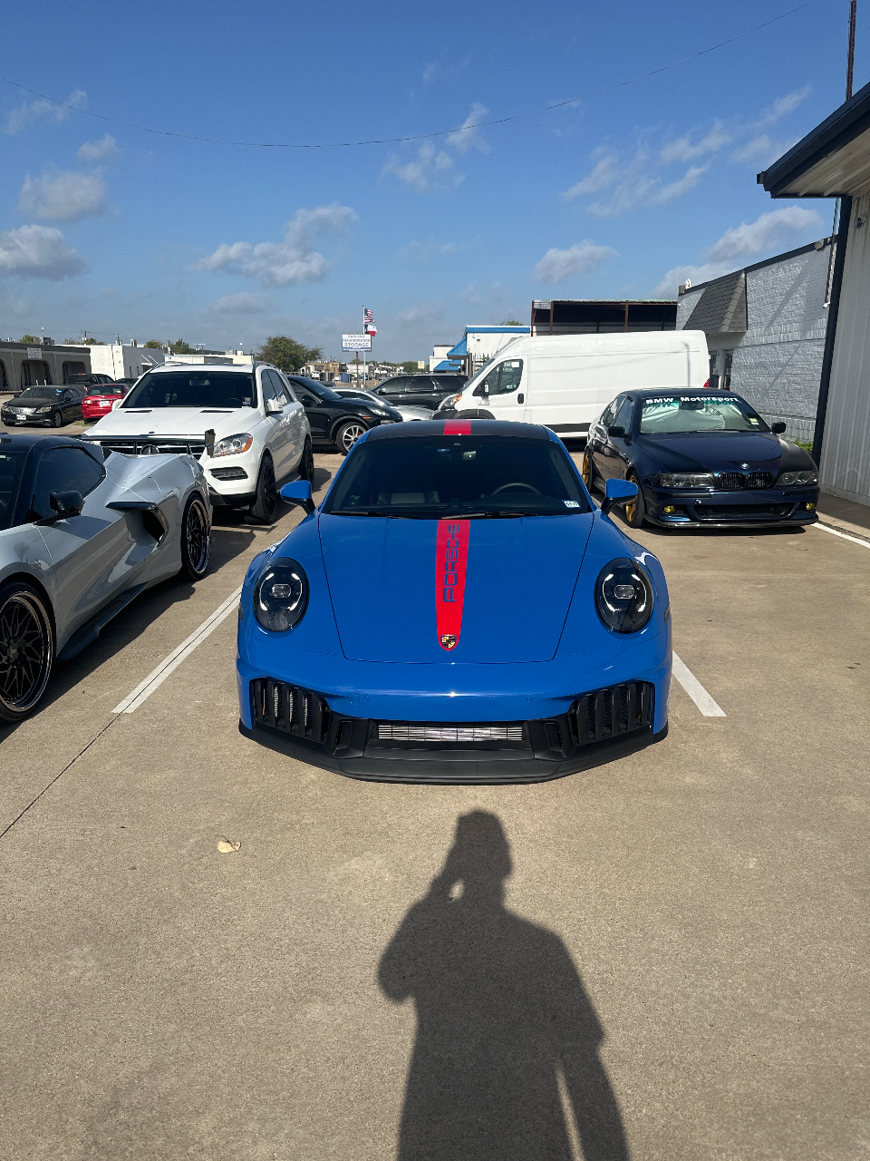 A blue Porsche 911 with a red racing stripe parked in a sunny outdoor lot next to other vehicles.