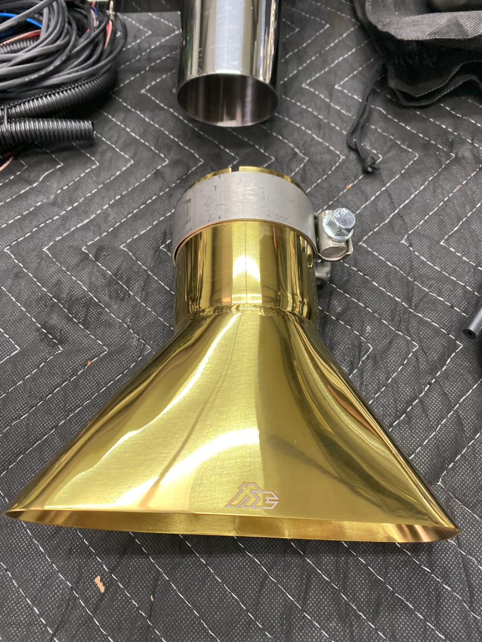 Close-up of a gold Jotech Motorsports exhaust tip with a polished finish and logo on a black quilted mat.