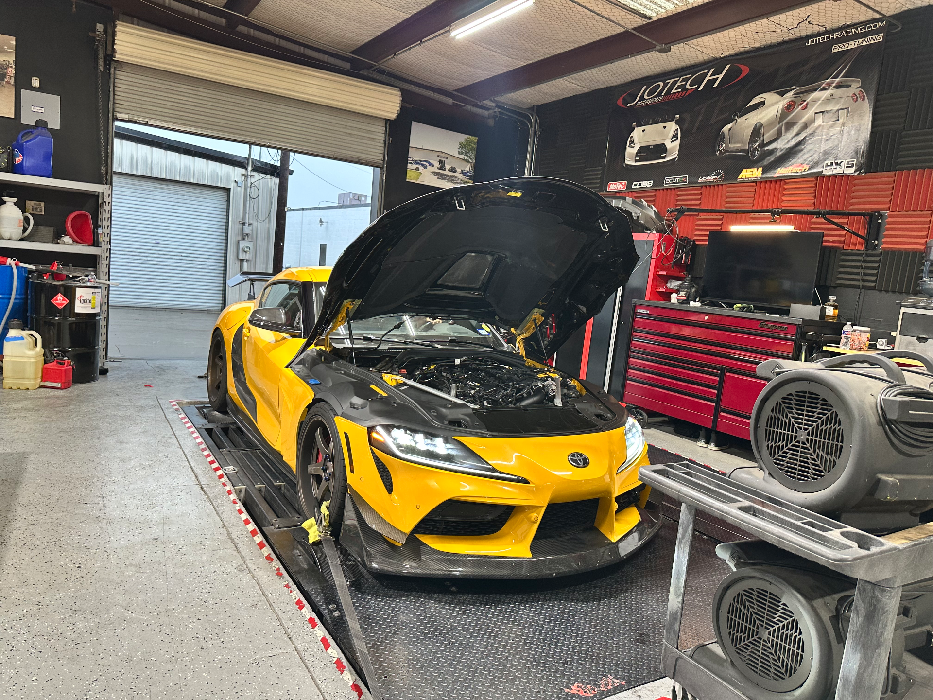 A yellow Toyota Supra with its hood open, strapped to a dynamometer in a Jotech Motorsports tuning shop.
