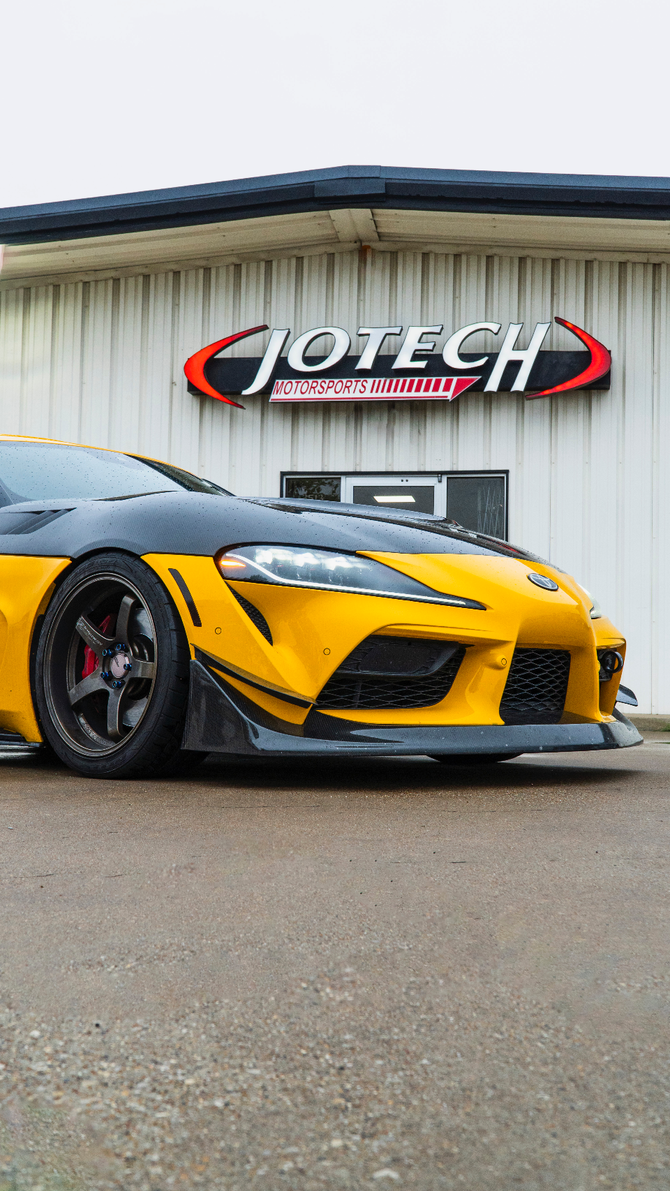 A yellow and black Toyota Supra with carbon fiber aero parked in front of the Jotech Motorsports shop.