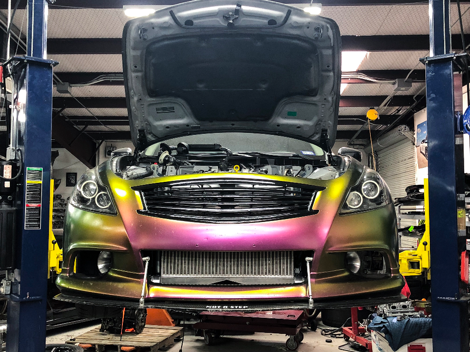 A chameleon-wrapped Infiniti G37 coupe on a lift in a performance shop, with its front bumper removed to reveal a large intercooler.