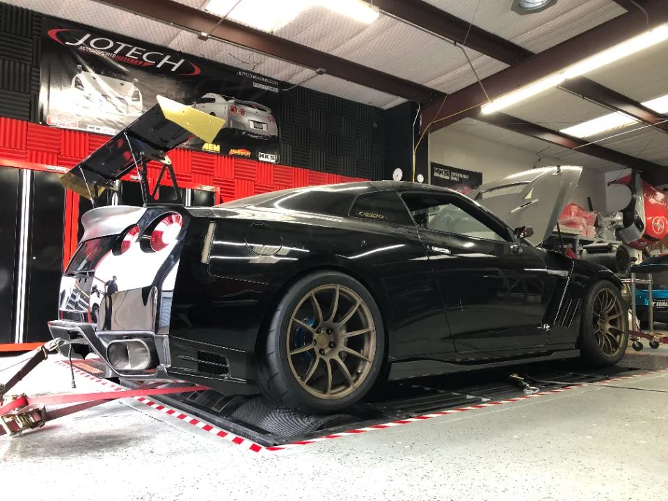 Black Nissan GT-R on a dyno at Jotech Motorsports with an aftermarket rear wing and bronze wheels, ready for tuning.