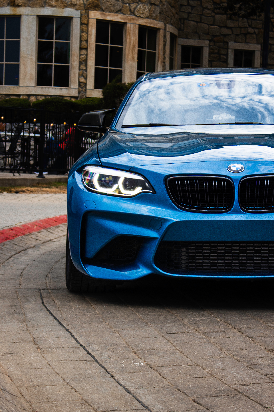 Front view of a vibrant blue BMW M2 parked on a paved surface in front of a stone building.