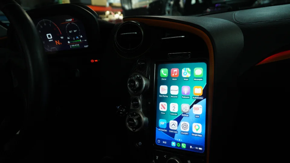 Interior view of a McLaren dashboard showing the digital instrument cluster and a vertically mounted smartphone displaying various apps.