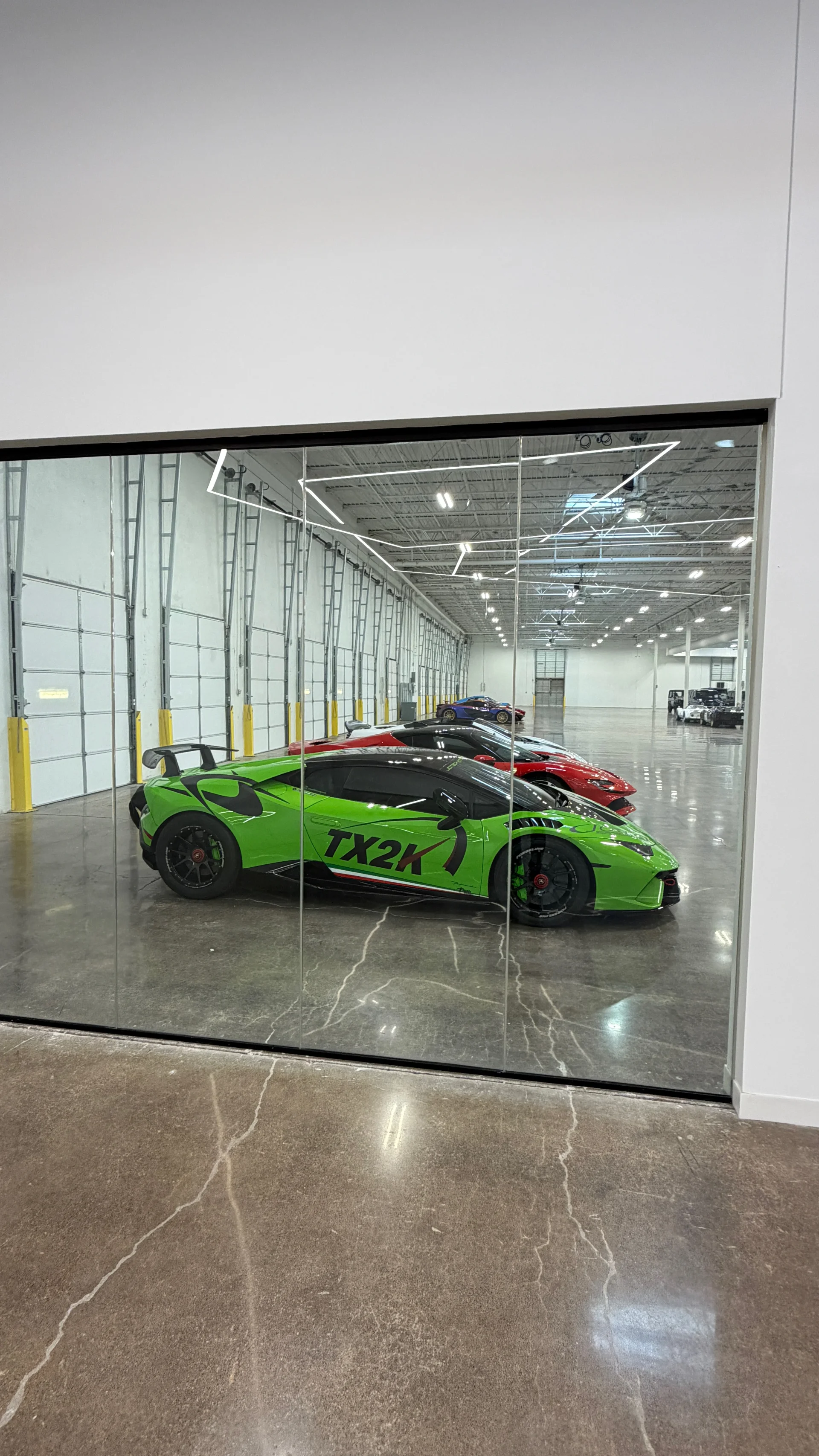 A line of high-performance Lamborghini supercars, including a green TX2K car, parked inside a large, well-lit facility.