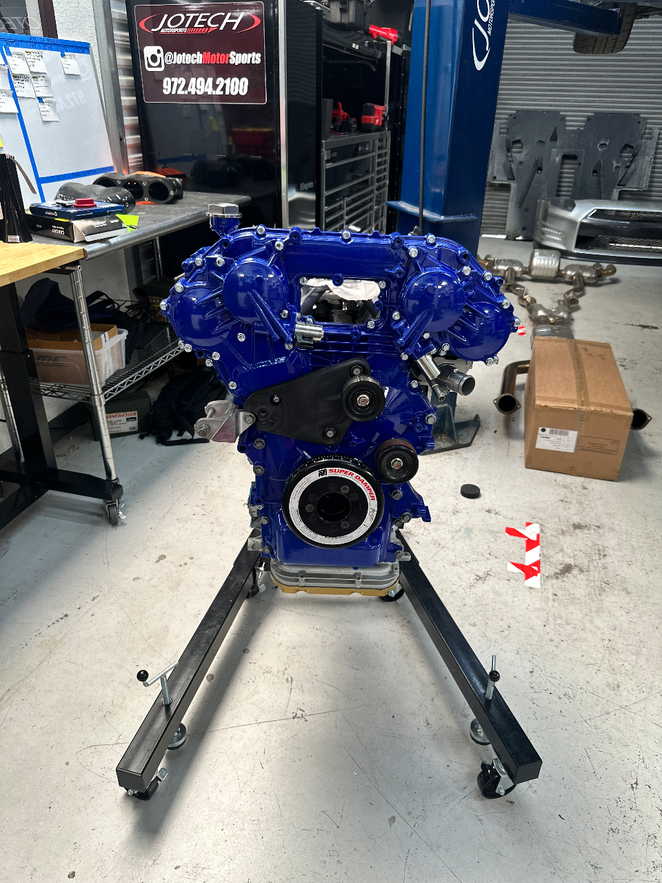 A custom blue built engine on an engine stand in the Jotech Motorsports shop, with tools and equipment in the background.