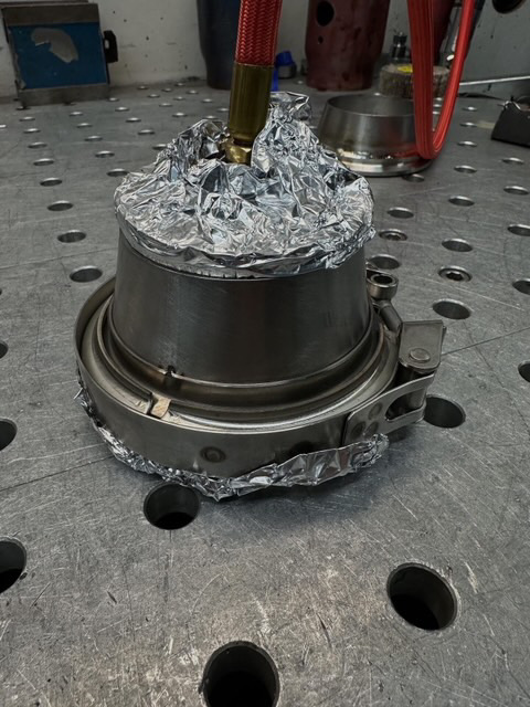 A metal turbo or exhaust component with a V-band clamp, partially covered in aluminum foil, on a shop workbench.