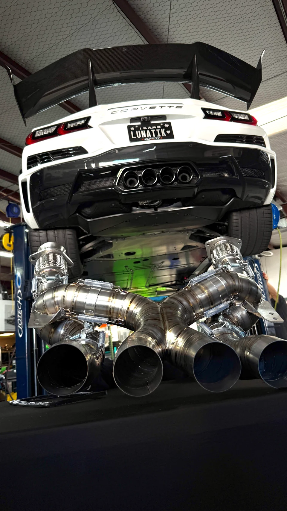 A white Corvette C8 on a lift in a shop, showcasing a custom, large-diameter aftermarket exhaust system with multiple pipes.