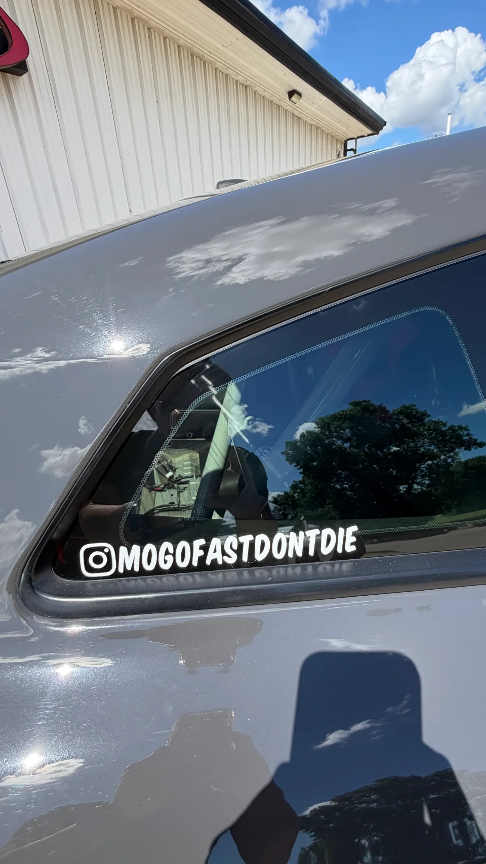 A close-up of a grey car's rear window featuring a white vinyl sticker with an Instagram handle that reads "MOGOFASTDONTDIE".