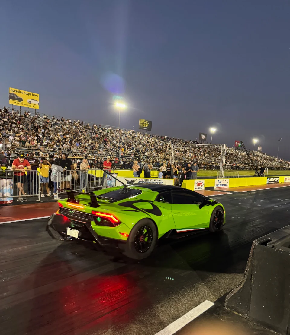 A bright green Lamborghini Huracan performs a launch at a drag strip at night, with spectators in the stands.