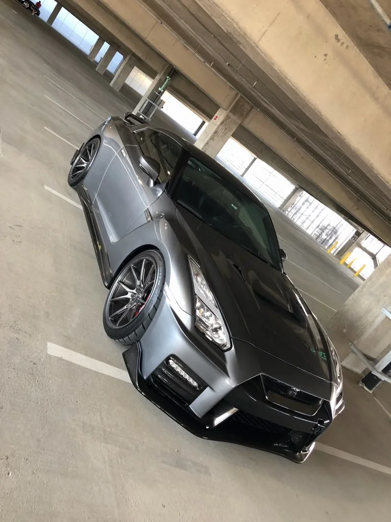 A grey Nissan GT-R R35 with aftermarket wheels and carbon fiber aerodynamic parts parked in a concrete parking garage.