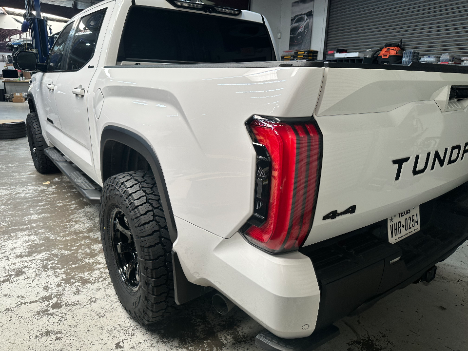 Rear view of a white Toyota Tundra pickup truck with black off-road wheels and tires in a garage setting.