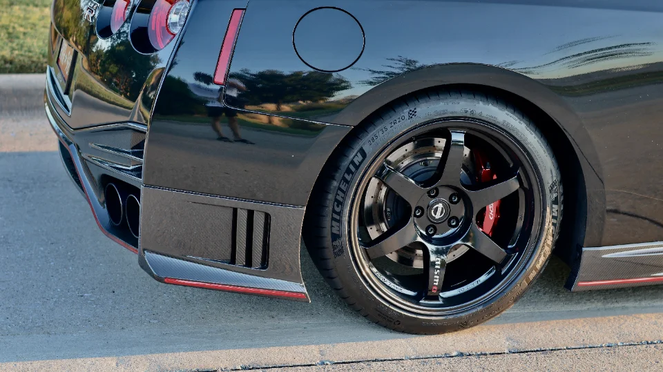 Close-up of a black Nissan GT-R's rear wheel, featuring black aftermarket wheels, Michelin tires, red Nismo brakes, and a carbon fiber rear diffuser.