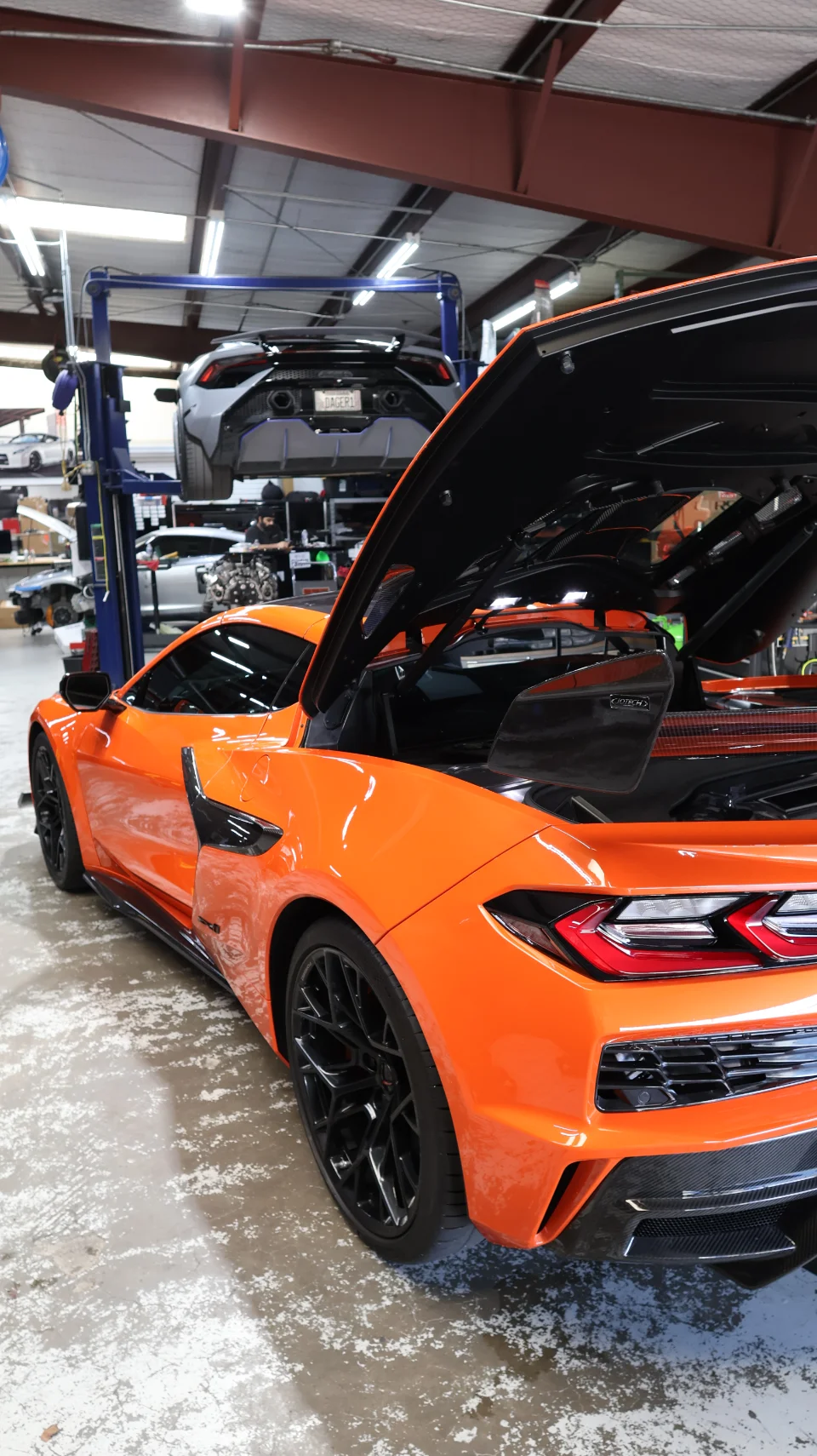 An orange Corvette C8 with its engine bay open in a performance tuning shop, with a Lamborghini Huracan on a lift in the background.