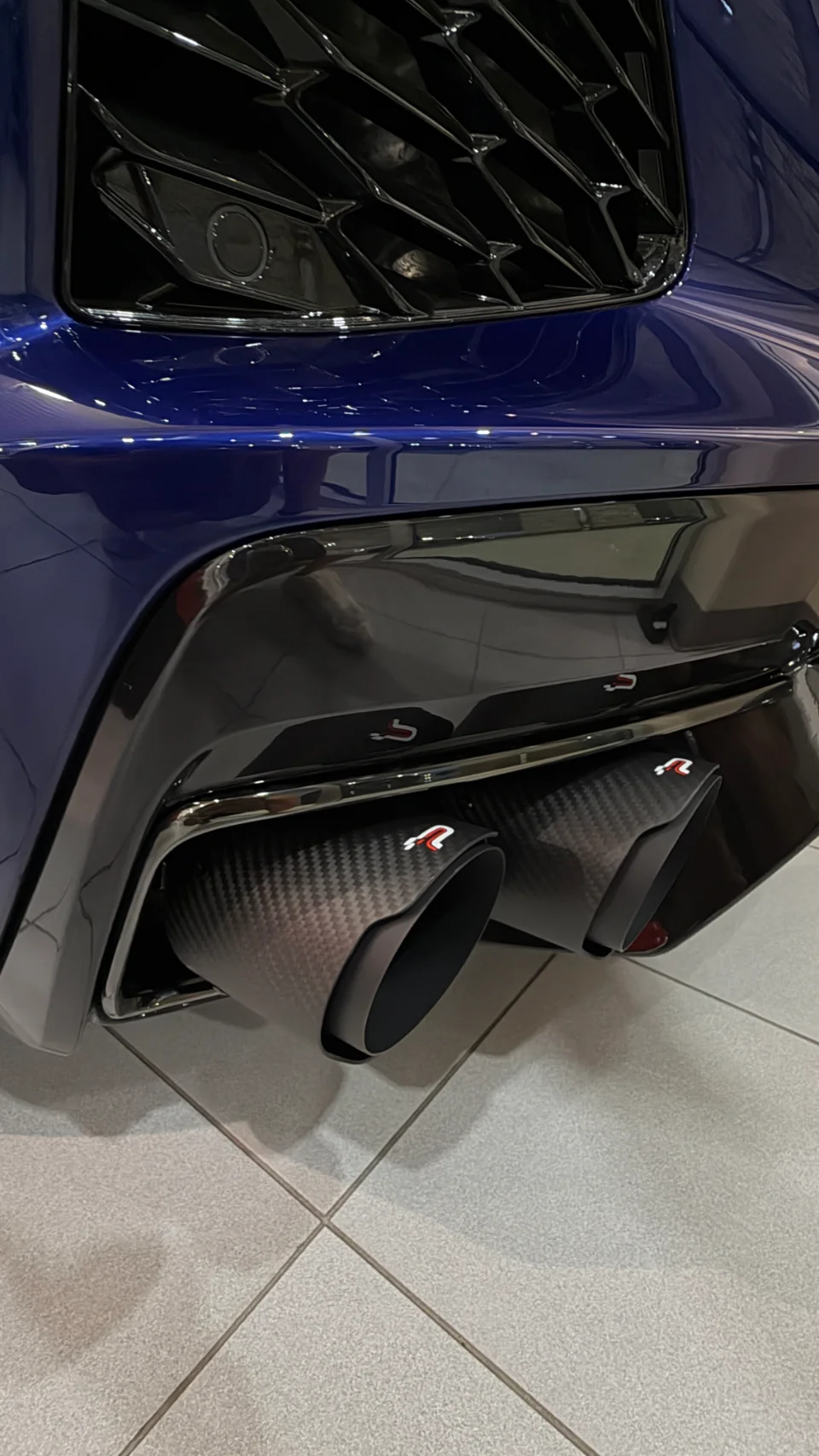 Close-up of a blue car's rear bumper featuring aftermarket carbon fiber exhaust tips with red logos.