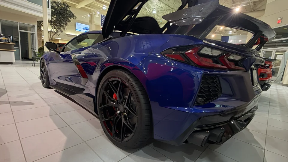 A blue Chevrolet Corvette C8 with aftermarket wheels and a carbon fiber rear wing displayed in a showroom.