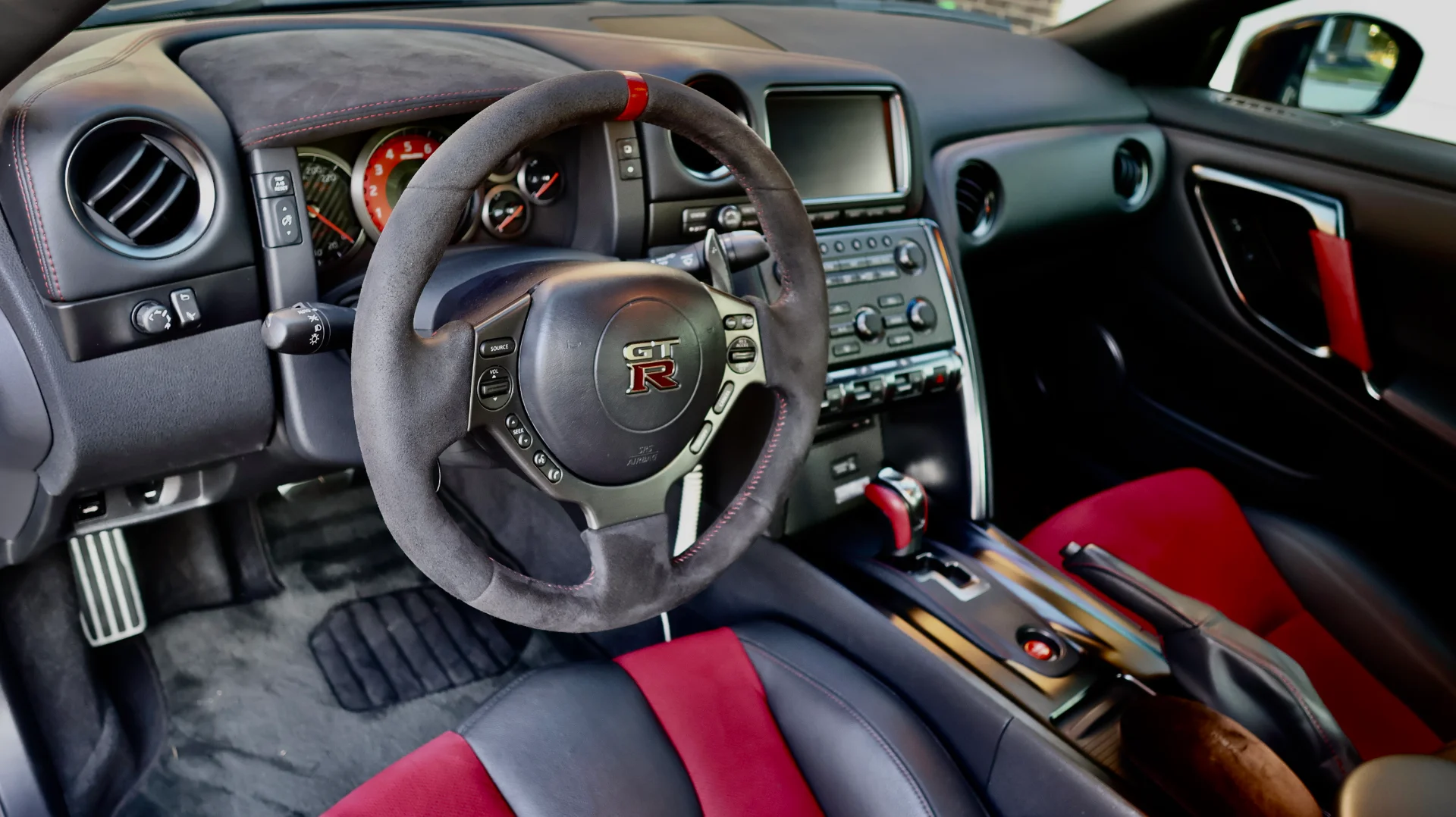 Detailed interior shot of a Nissan GT-R featuring an alcantara steering wheel, red and black seats, and dashboard.