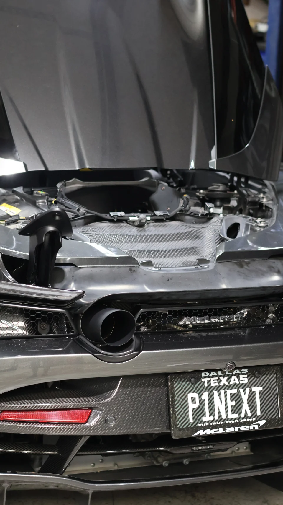 Rear view of a grey McLaren in a shop with its engine bay open, showcasing a large exhaust tip and carbon fiber details.