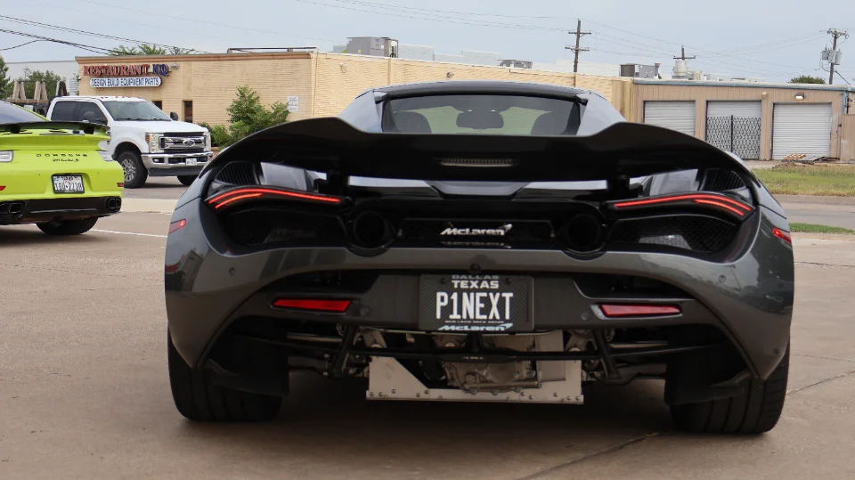 Rear view of a grey McLaren 720S with a custom license plate, parked on a street next to a green Porsche 911 GT3 RS.