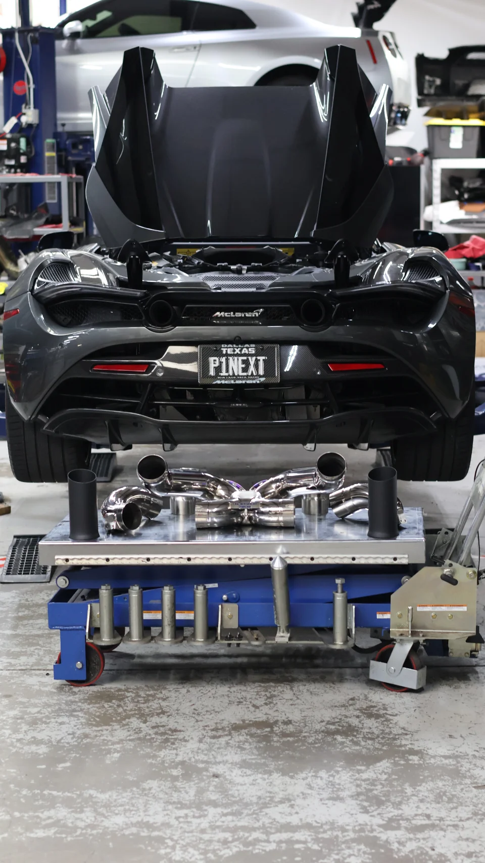 Rear view of a grey McLaren on a lift in a shop, with a performance exhaust system displayed on a cart below.