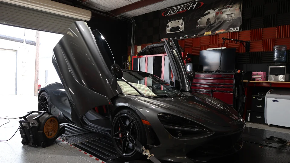 A grey McLaren 720S supercar with its butterfly doors open, strapped to a dynamometer in a performance tuning shop.