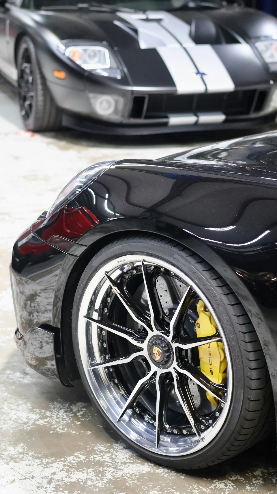 Close-up of a black Porsche's custom wheels and yellow brake calipers, with a black Ford GT in the background.