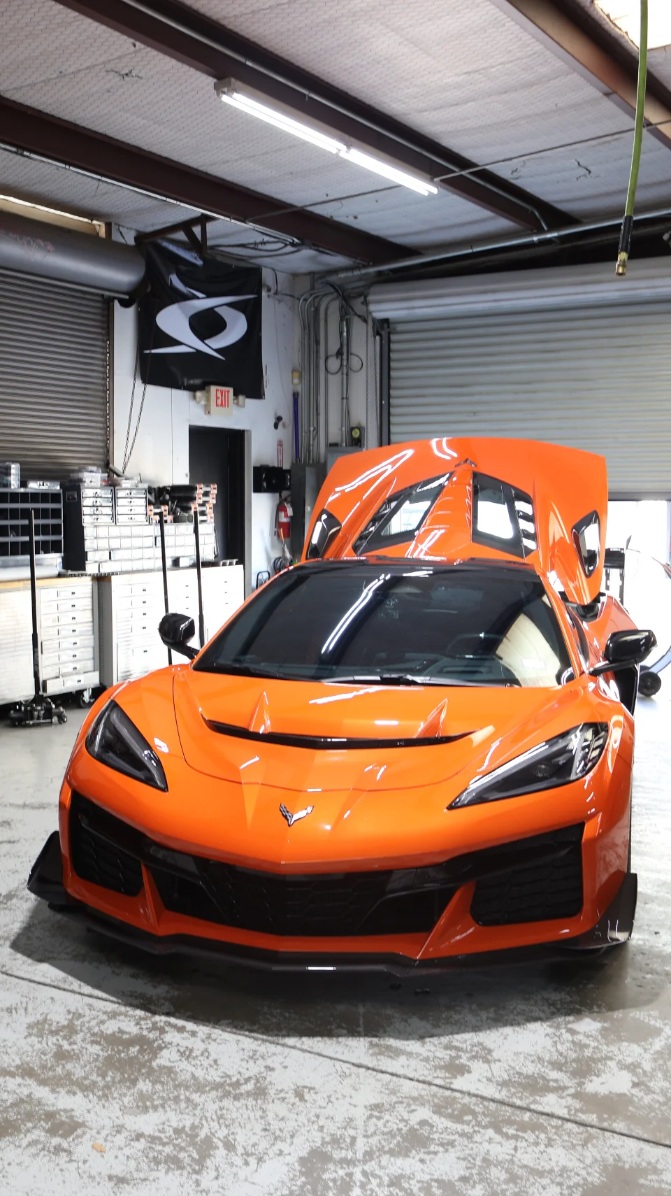 An orange Chevrolet Corvette C8 Z06 with its engine bay open, parked inside a performance tuning shop.