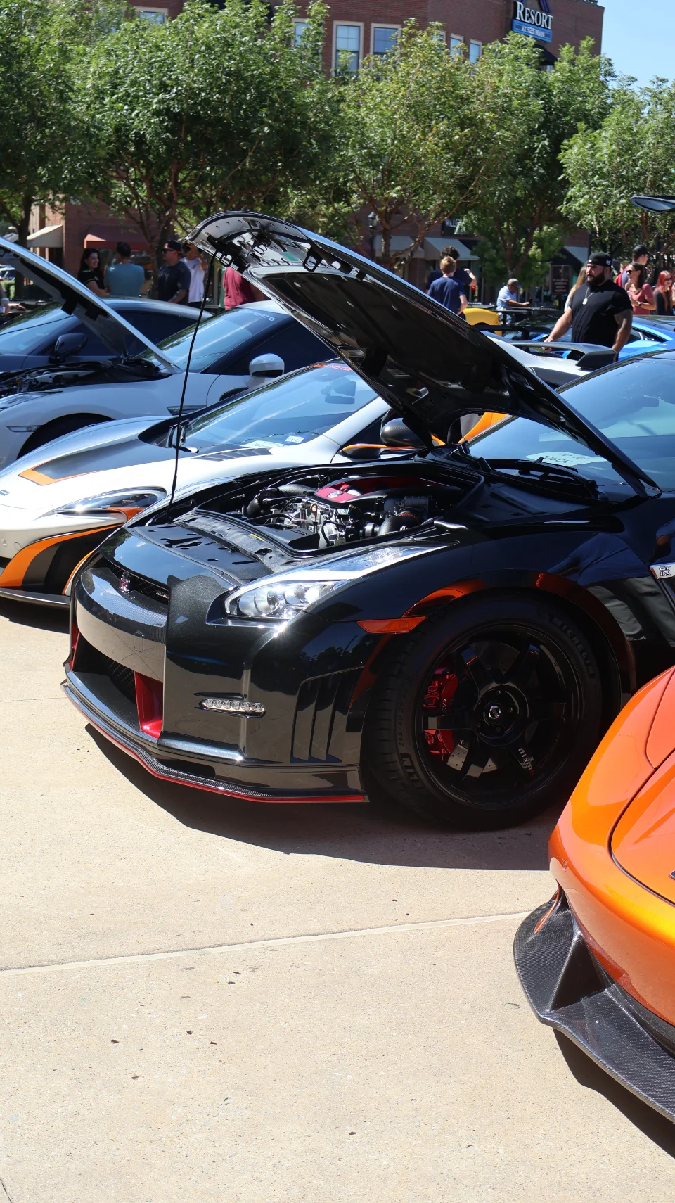 A black Nissan GT-R with its hood open, showcasing the engine bay, parked next to a white McLaren at a car show.