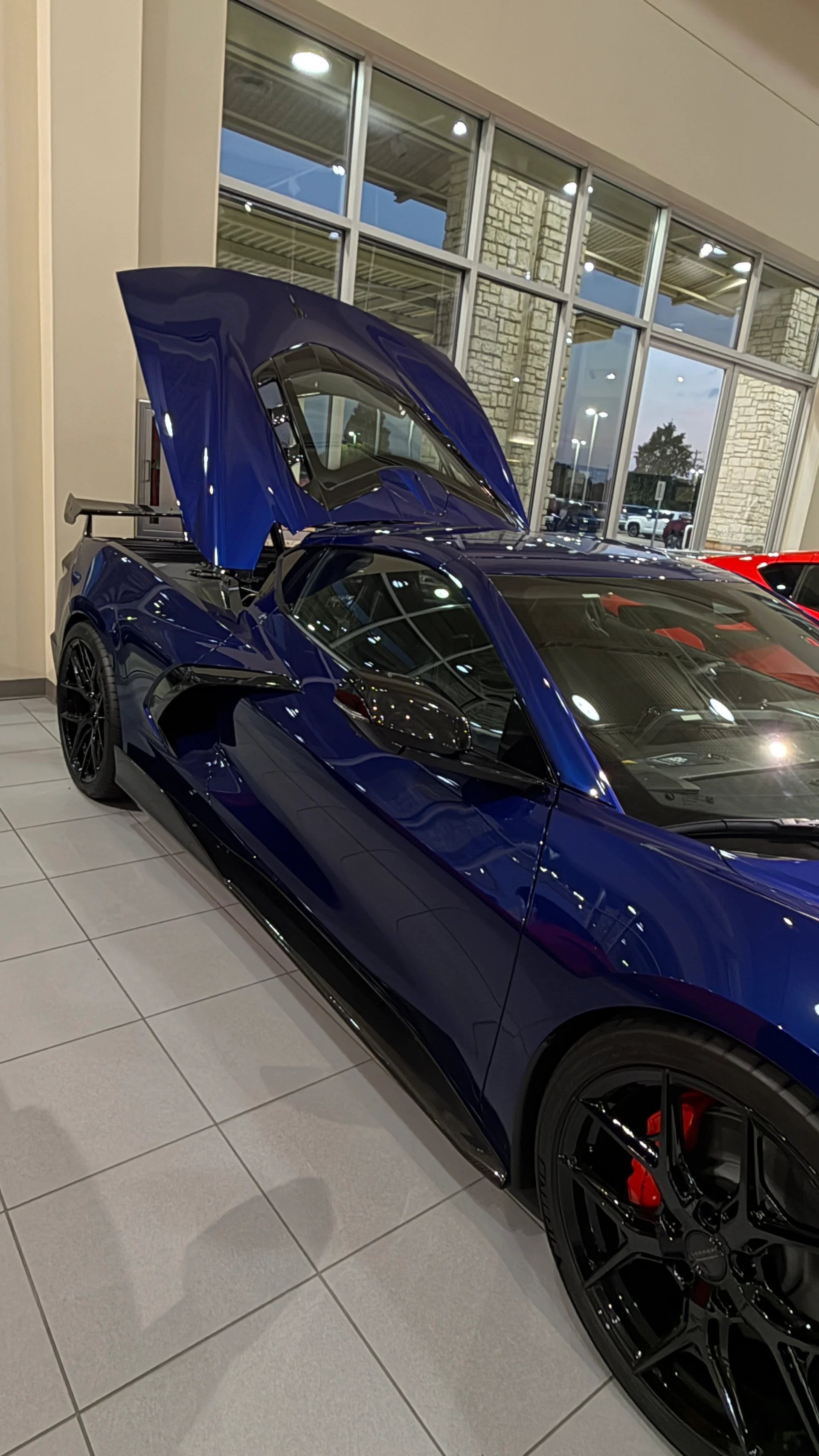 A blue Chevrolet Corvette C8 with its engine cover open, displayed in a car dealership showroom.