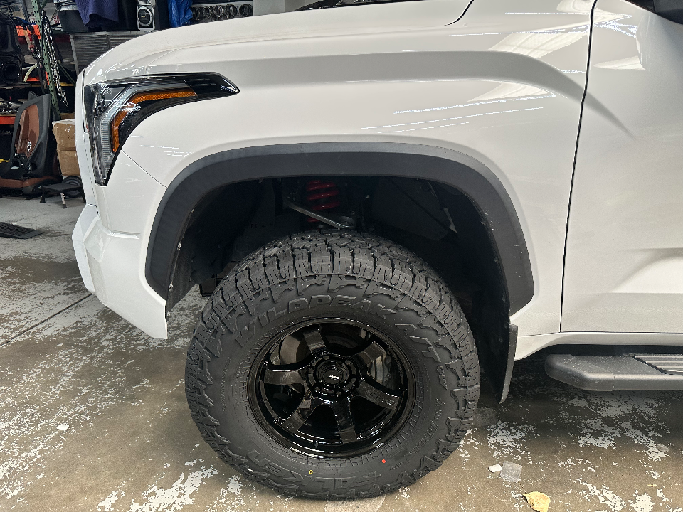 Close-up of a white truck's front wheel with black off-road tires and visible red suspension coil in a shop.