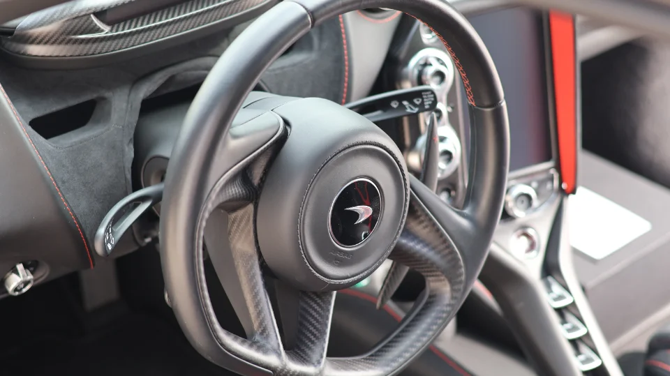 Close-up of a McLaren interior, showing the carbon fiber and leather steering wheel with red stitching and dashboard.