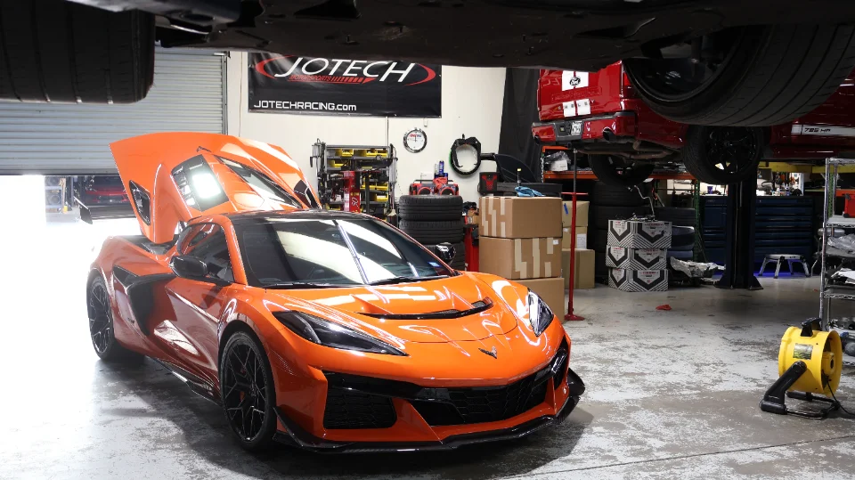 An orange Chevrolet Corvette C8 Z06 with its engine cover open, parked inside the Jotech Motorsports shop.