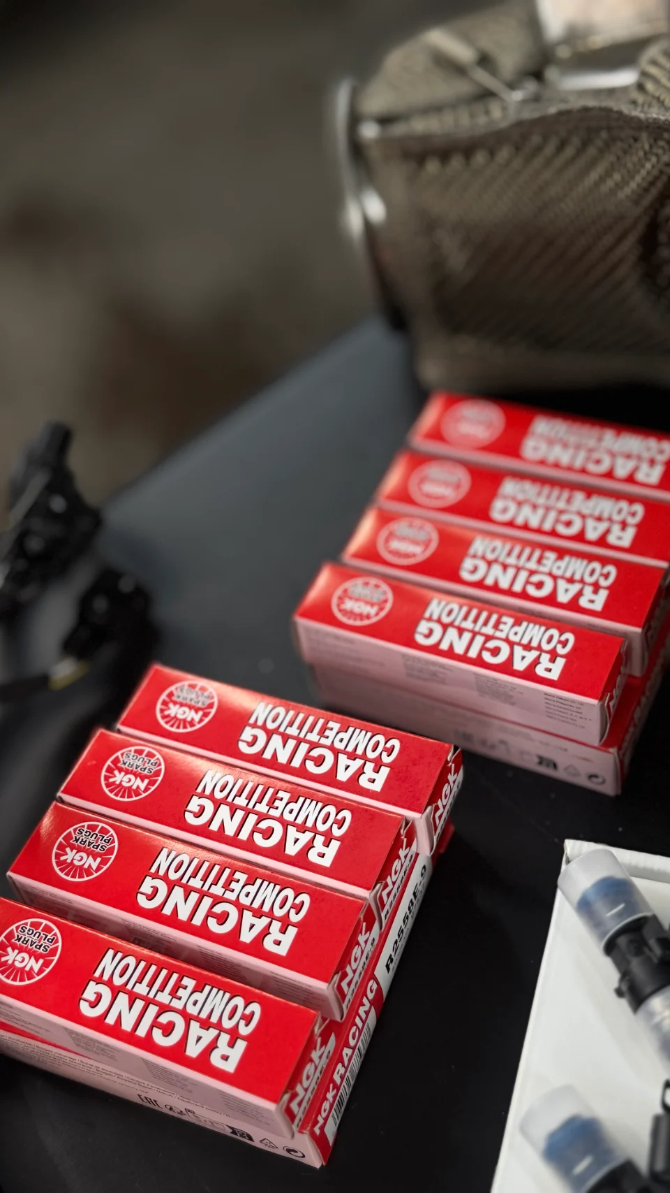 Boxes of NGK Racing Competition spark plugs on a dark surface, ready for installation in a performance vehicle.