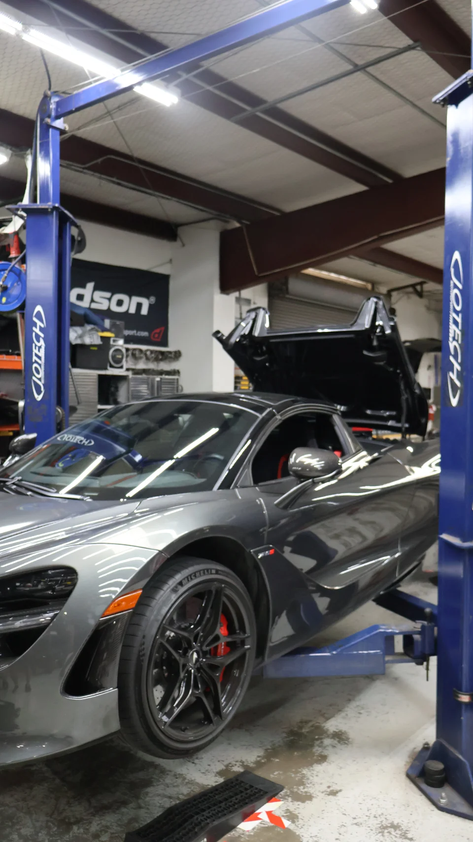 A grey McLaren 720S on a blue hydraulic lift in a Jotech Motorsports performance tuning shop.