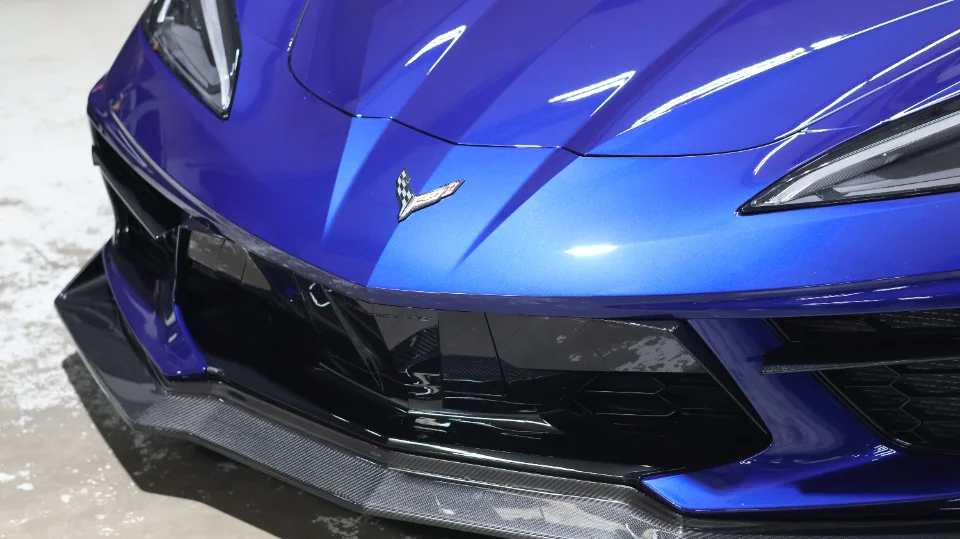 A close-up of the front of a blue Chevrolet Corvette C8, featuring a carbon fiber front splitter and the Corvette emblem.