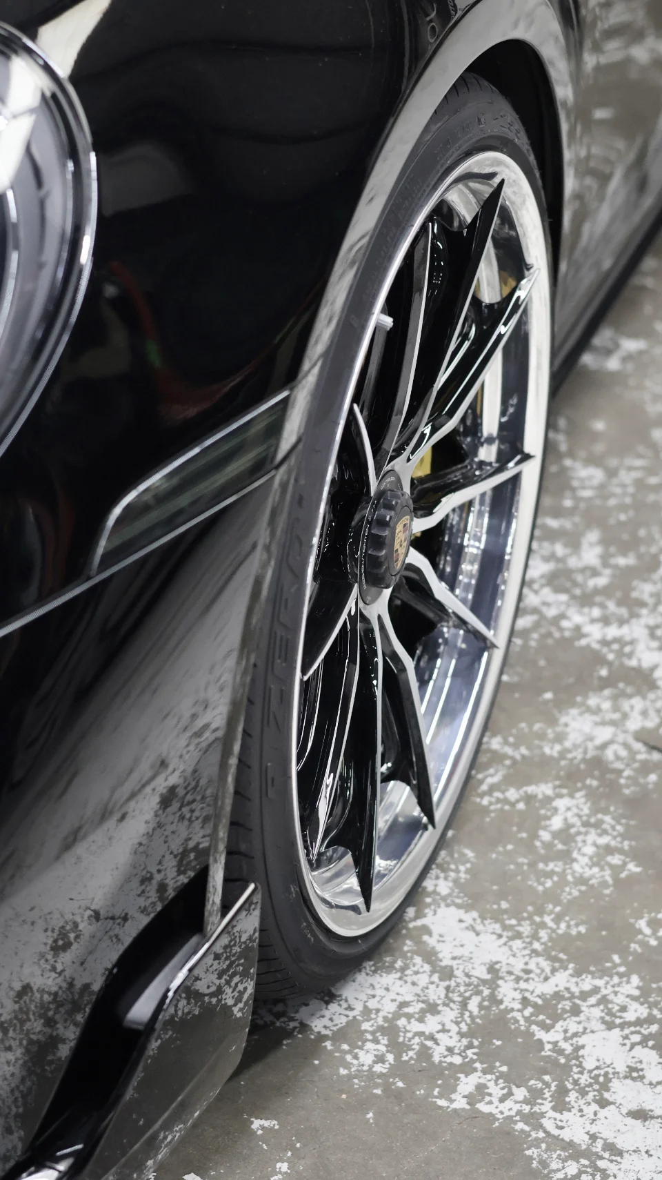 Close-up detail of a black Porsche's chrome multi-spoke wheel and tire on a concrete floor.