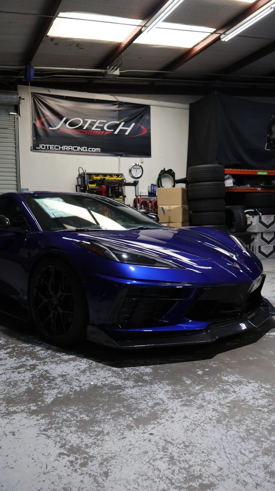 Blue Chevrolet Corvette C8 with a carbon fiber front splitter parked inside the Jotech Motorsports shop.