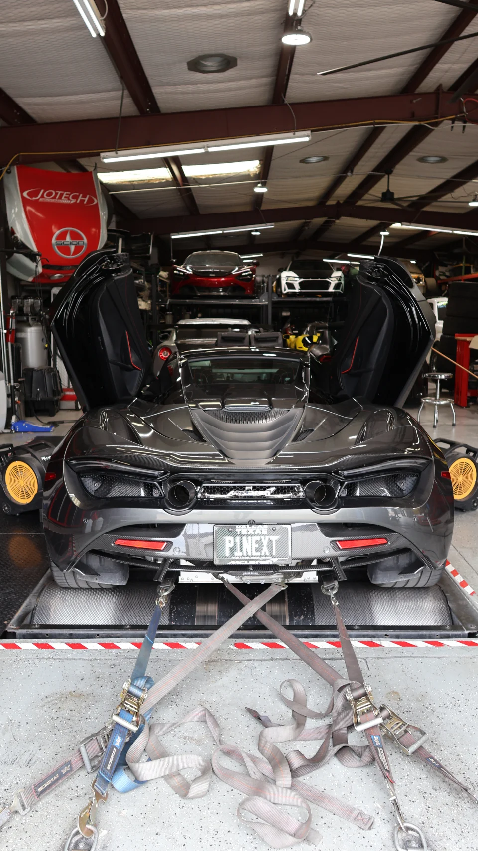 A black McLaren 720S with its butterfly doors open, strapped to a dyno in a performance tuning shop.