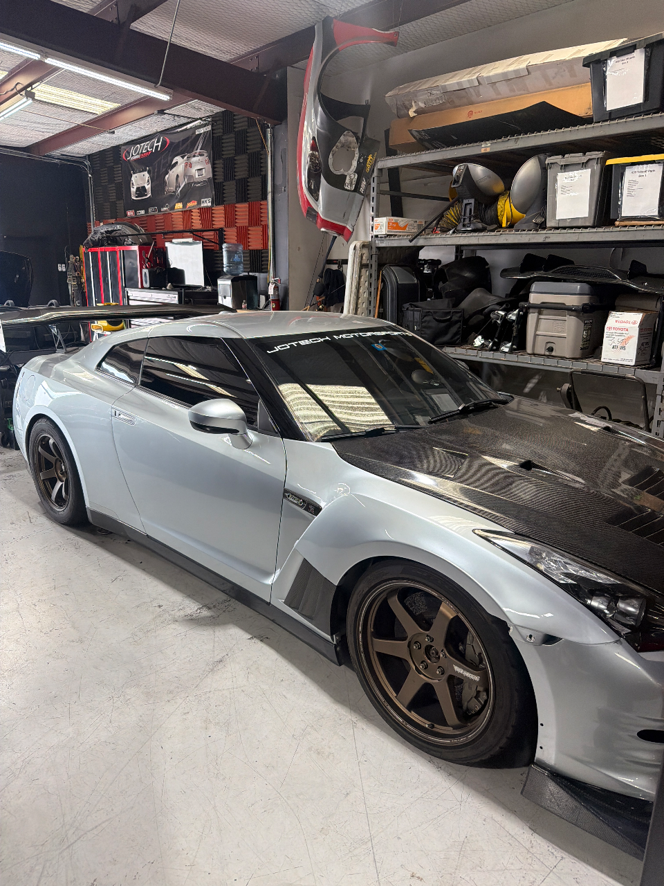 A silver Nissan GT-R with a carbon fiber hood and widebody kit parked inside the Jotech Motorsports shop.